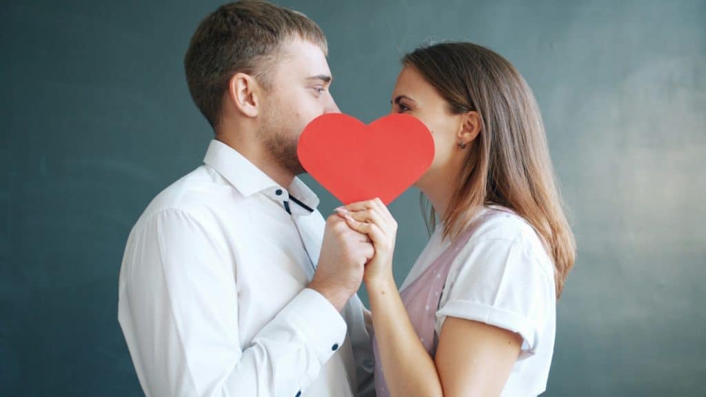 A couple in white shirts holding a large red paper heart between them.