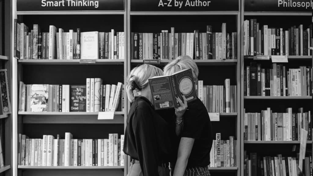 A boy and a girl standing in a school library hiding their faces with a book.