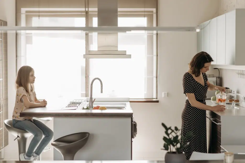 Girl Sitting at the Table in a Kitchen and Woman Pouring Juice into a Glass