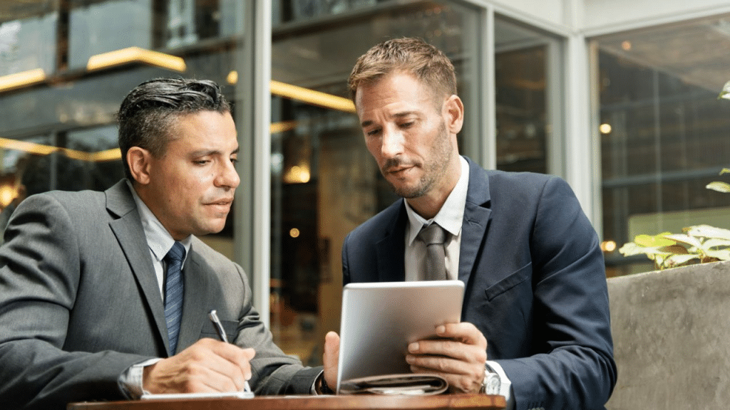 Two businessmen in suits look intently at a tablet while one writes on a notepad.