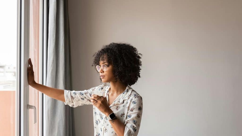 A woman standing by a window holding a cup, looking outside thoughtfully.