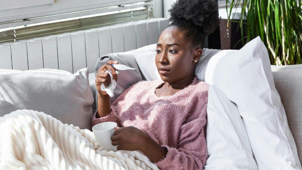 A woman lying on a couch with a blanket, holding a tissue and a cup.