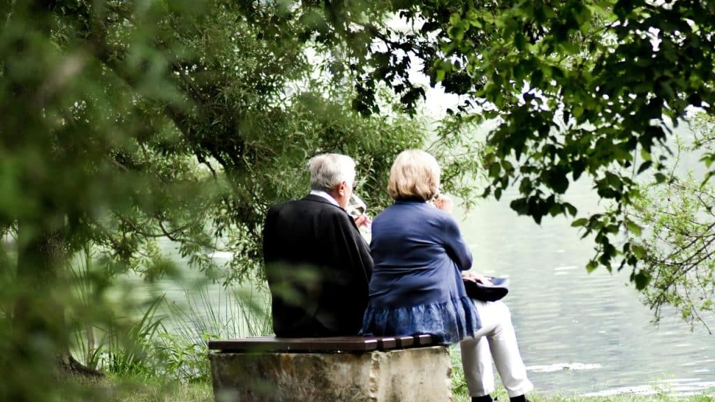 A senior couple sitting on a bench by a lake, surrounded by trees and greenery.
