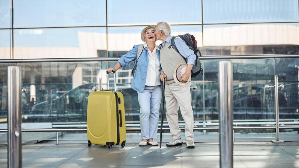 A senior couple at an airport, smiling and sharing a kiss while holding luggage and travel bags.