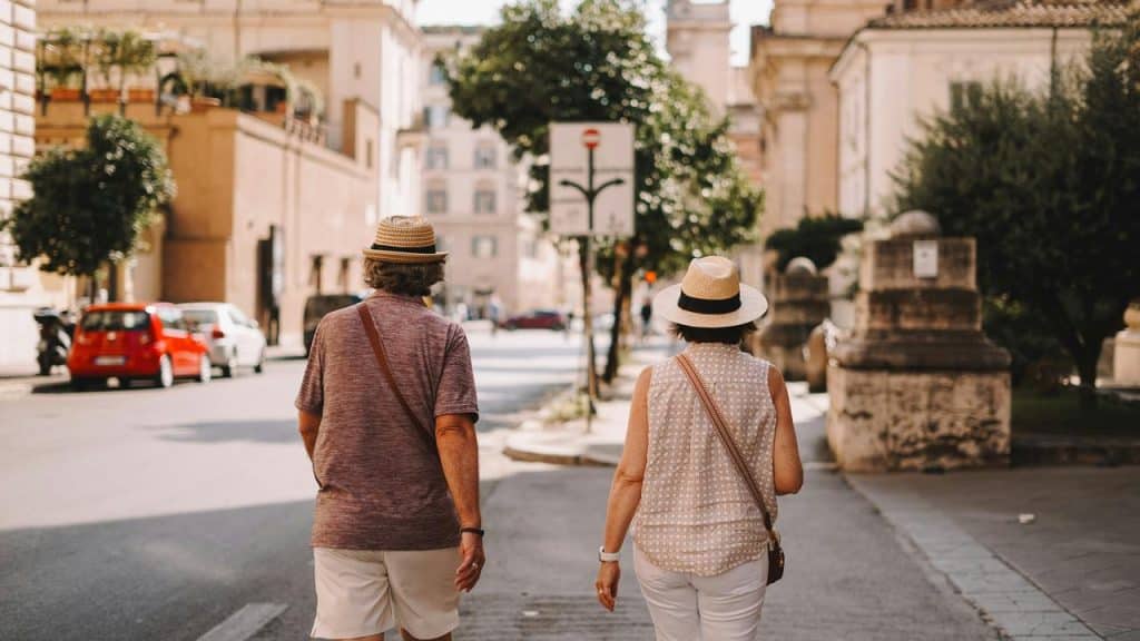 A senior couple walking down a sunny city street wearing summer clothes and straw hats.