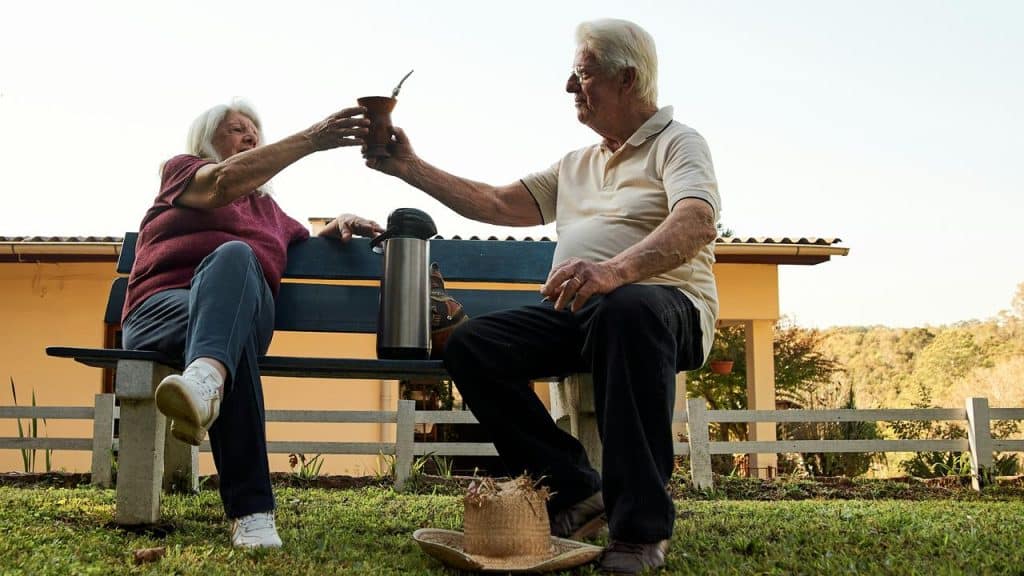 An elderly couple sitting on a bench sharing a drink outdoors.