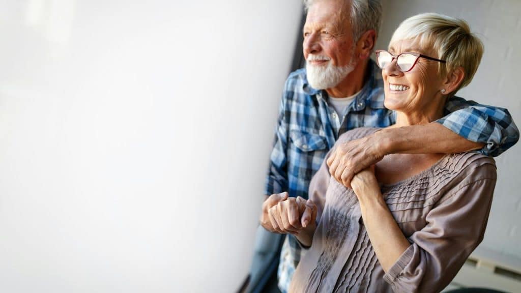 An elderly couple smiling and embracing while looking out a window.