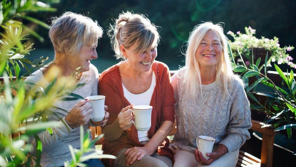 A group of older women sitting outdoors, laughing and holding coffee mugs.
