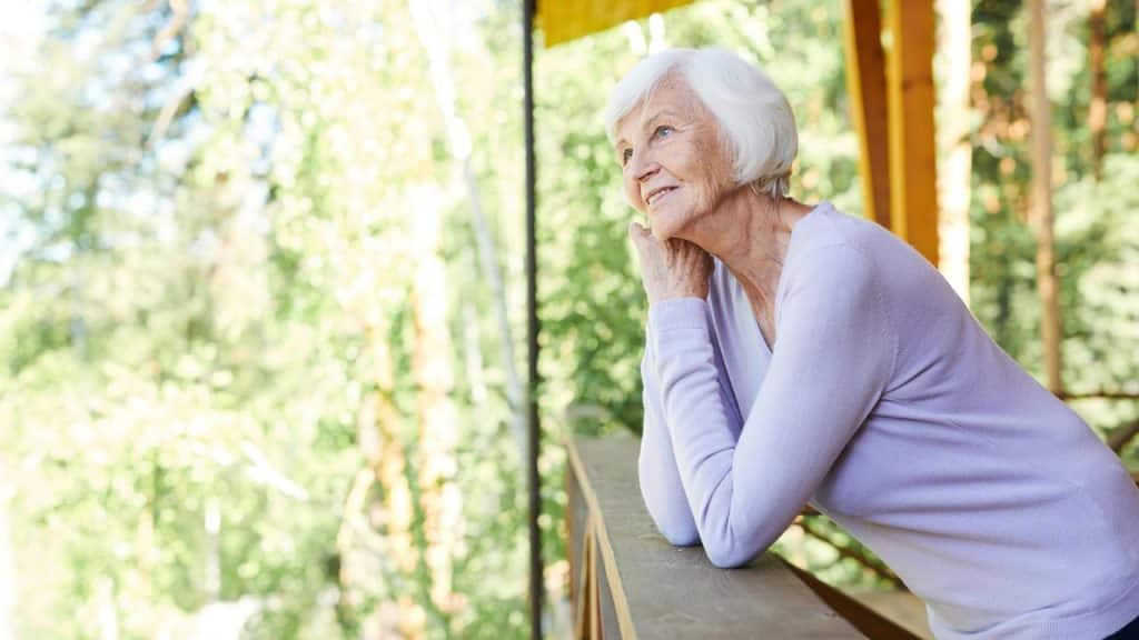 An elderly woman leaning on a balcony railing and smiling thoughtfully.