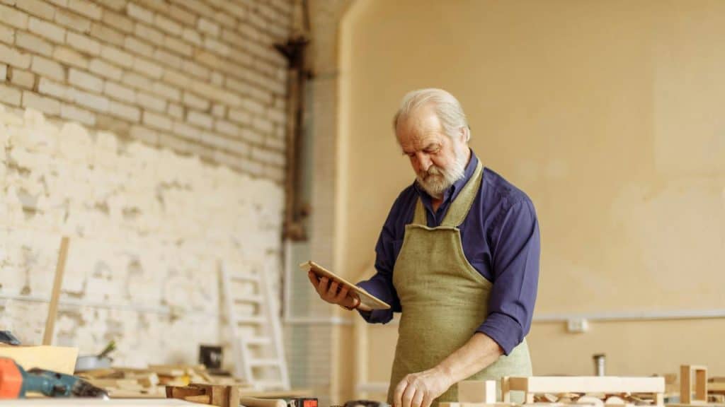 An elderly man wearing an apron working with wood in a workshop.