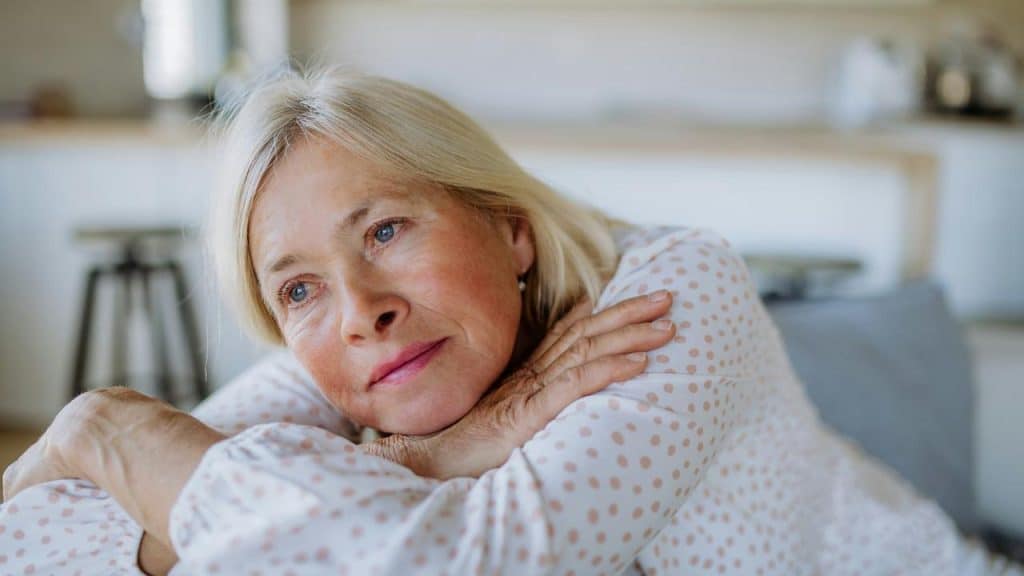 An elderly woman resting her head on her arms and looking thoughtful.