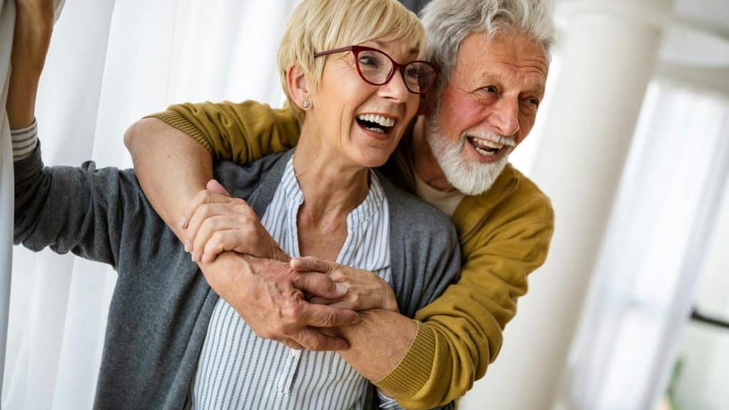 An elderly couple laughing and hugging near a window.