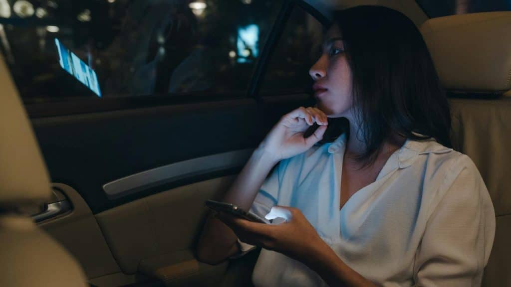 A woman sitting in the backseat of a car looking out the window while holding a phone.