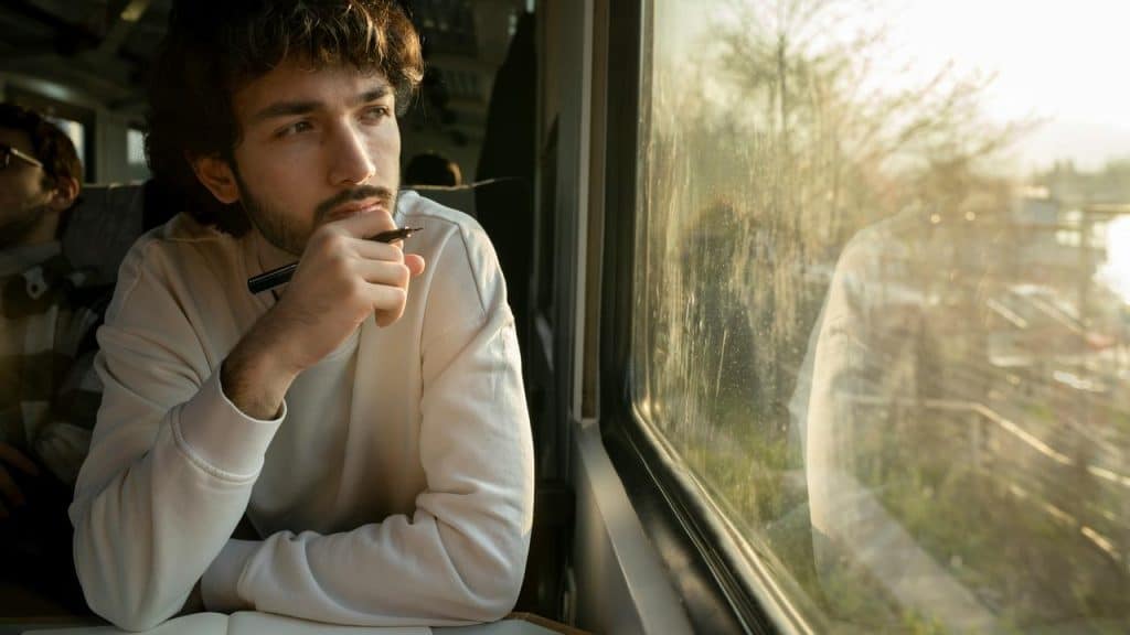 A man sitting by a train window holding a pen and looking outside thoughtfully.