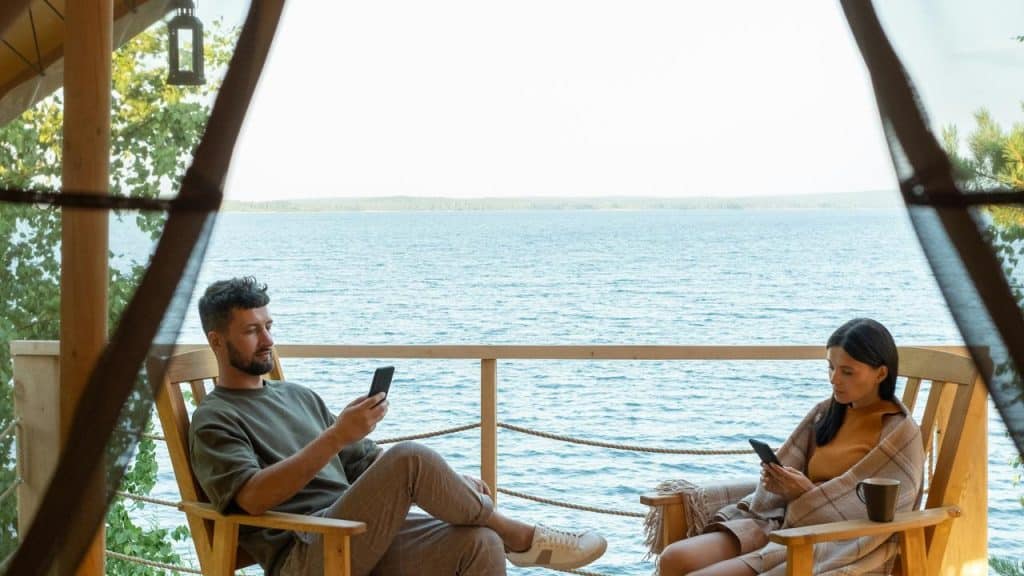A man and woman sitting on a deck by the water looking at their phones.