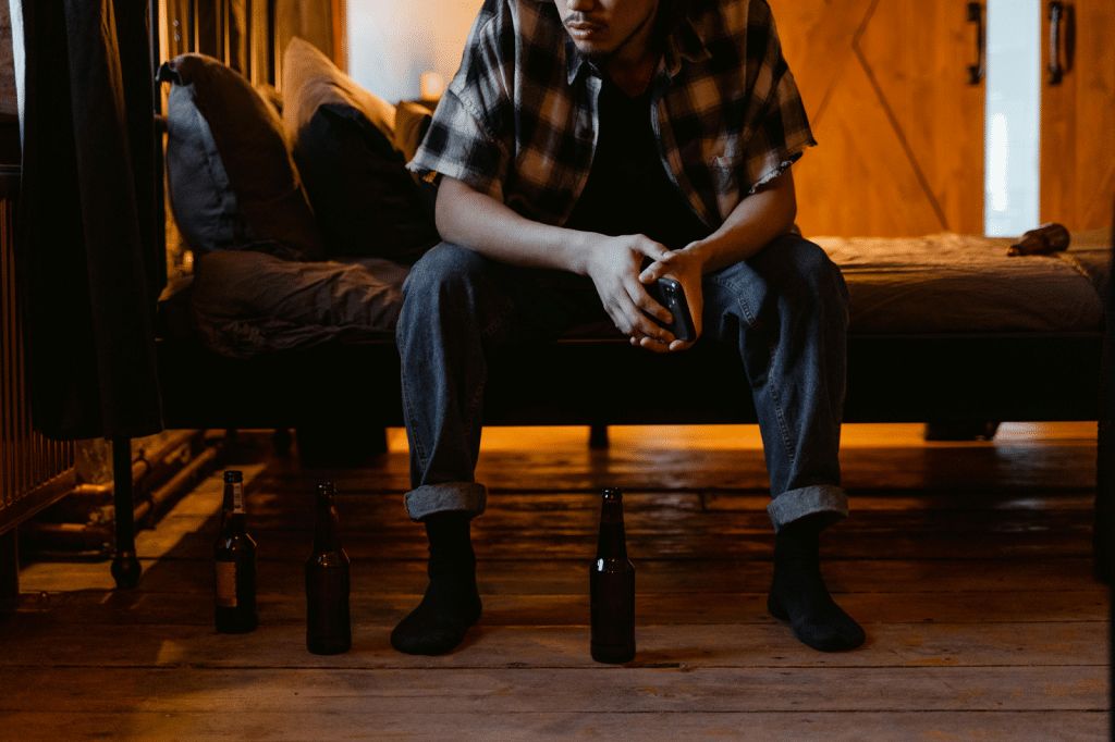 Man Holding a Cellphone and Sitting on a Bed with Beer Bottles on the Floor
