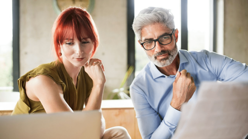 A younger woman and an older man with glasses look down at a laptop screen together.