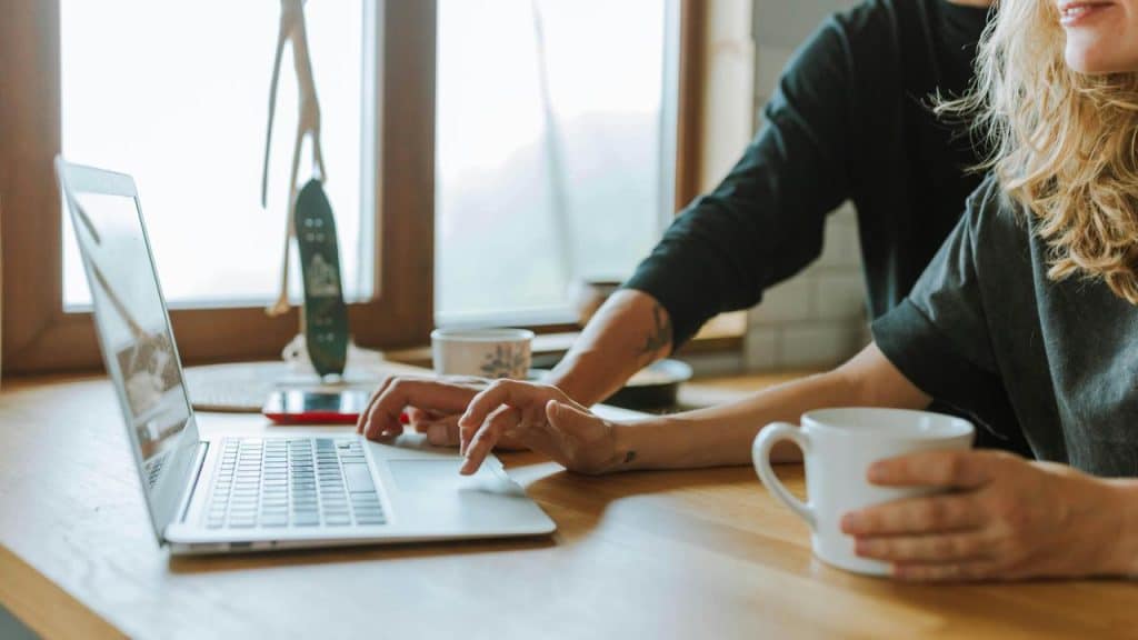 A person holding a mug while working on a laptop with someone pointing at the screen.