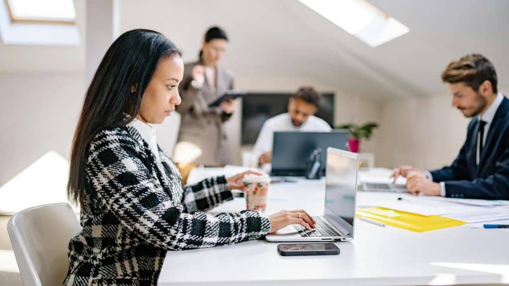 A woman working on a laptop at a table with colleagues in the background.