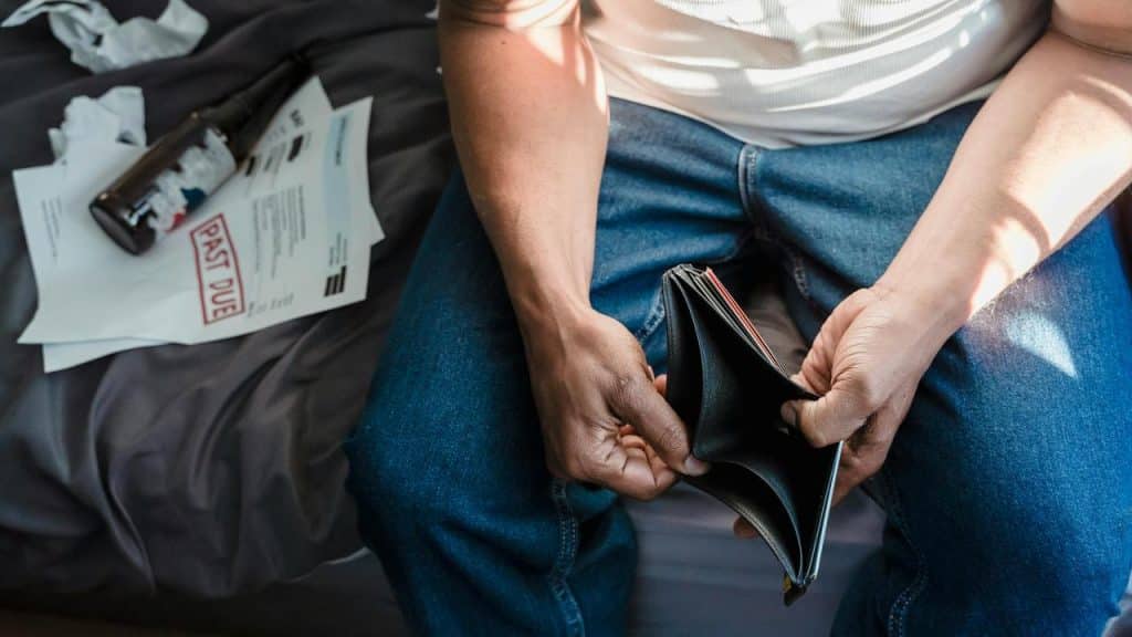 A man sitting on a bed holding an empty wallet beside overdue bills.