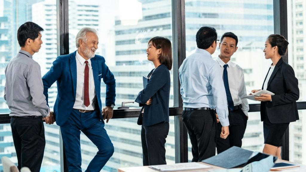 Six diverse professionals stand in two groups, talking near large office windows.