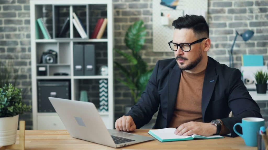 Man in a suit jacket and glasses looking at a laptop and writing in a notebook at a desk.