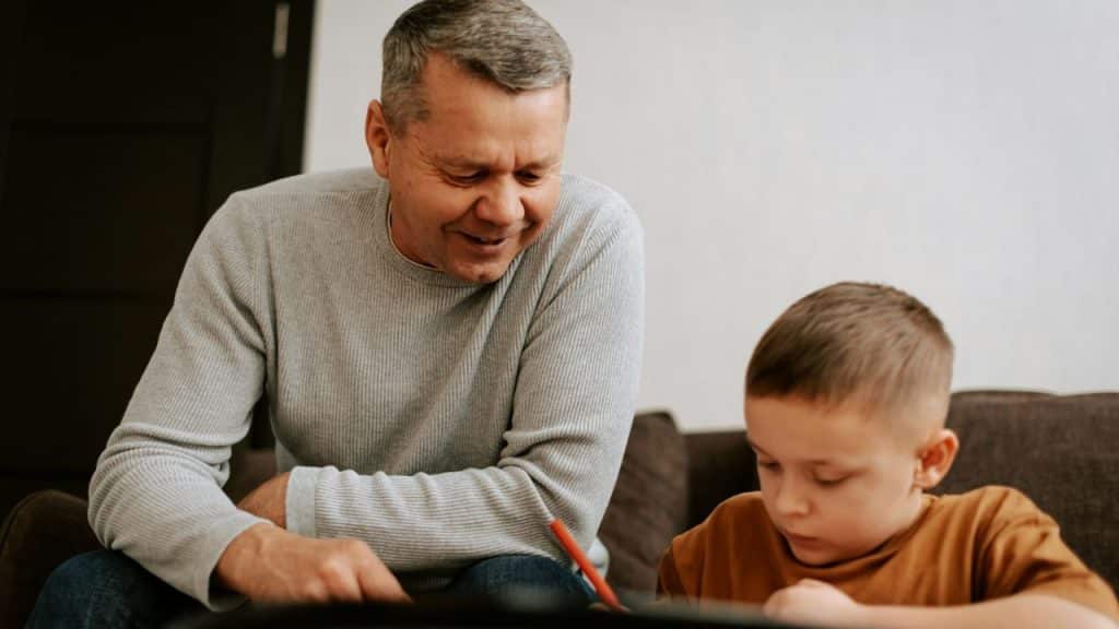 Smiling older man in a gray sweater watching a young boy write with a red pencil.