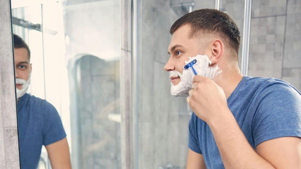 Man with shaving cream on his face shaving with a razor in front of a mirror.