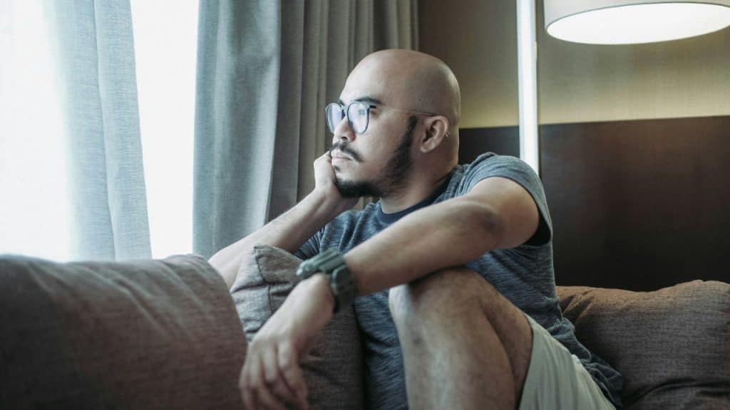 Bald man with a beard and glasses sitting on a couch, looking out a window.