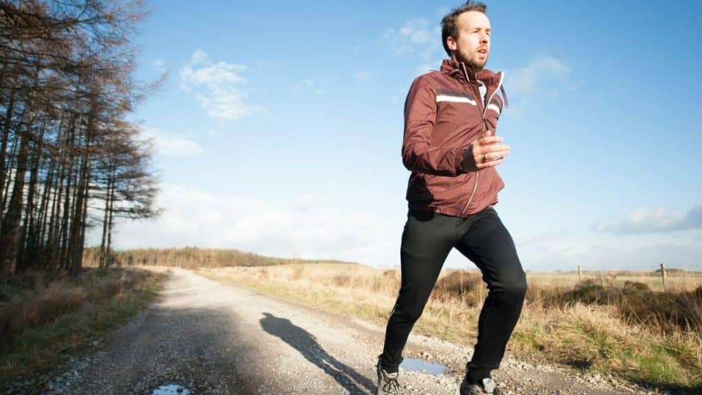 Man in a brown jacket and black pants jogging on a dirt road next to a field.