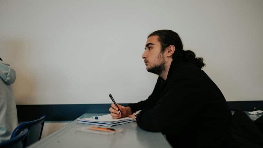 Young man with long hair in a ponytail sitting at a desk and writing with a pen.