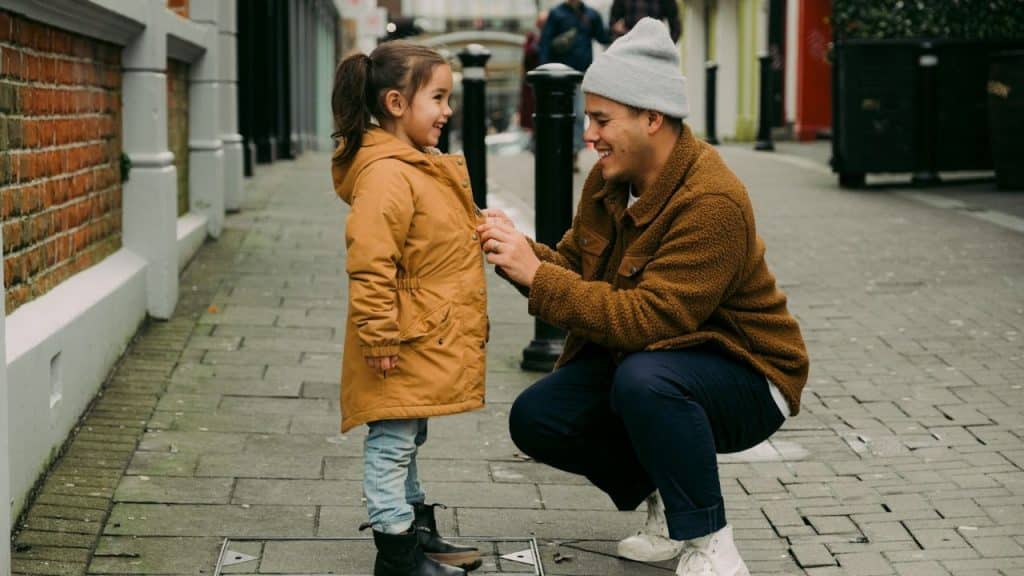 Man in a brown fleece jacket and beanie kneeling to button a young girl's mustard-yellow jacket.