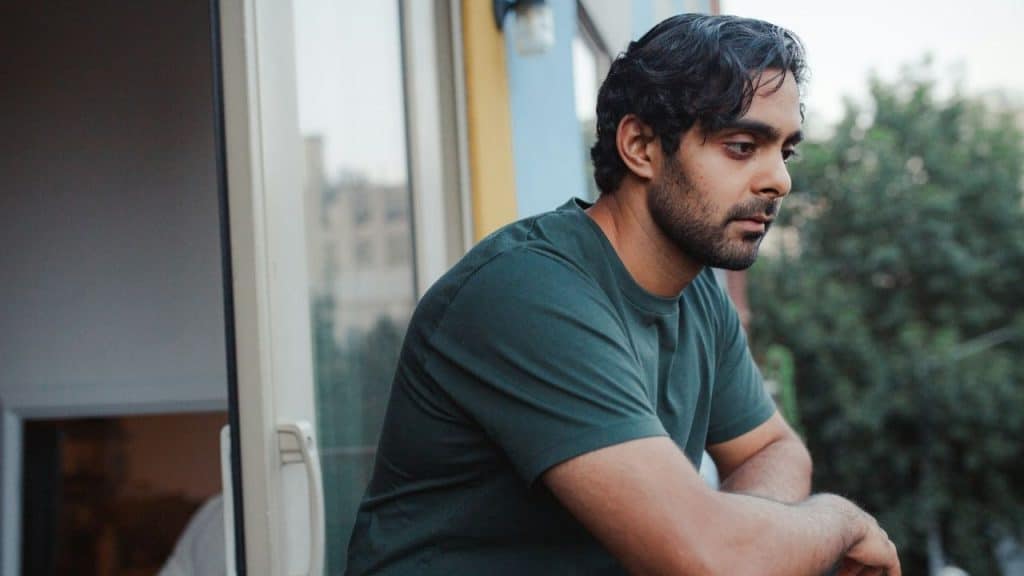 Man with dark hair and a beard standing pensively by a window looking out at trees.