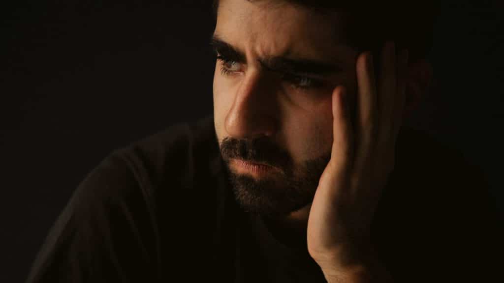 A man with a beard sits in darkness, his hand resting on his cheek, looking distressed.