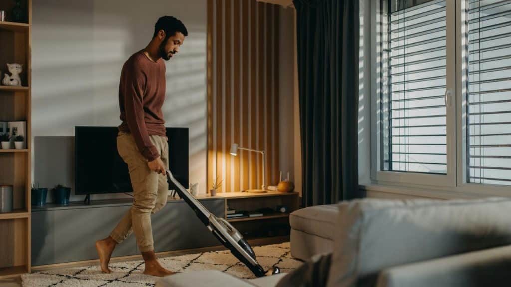 A barefoot man in a maroon sweater vacuums a shaggy white rug in a sunny living room.
