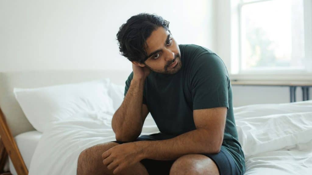 A young man sits on a white bed in a bright room, his hand resting on his neck.
