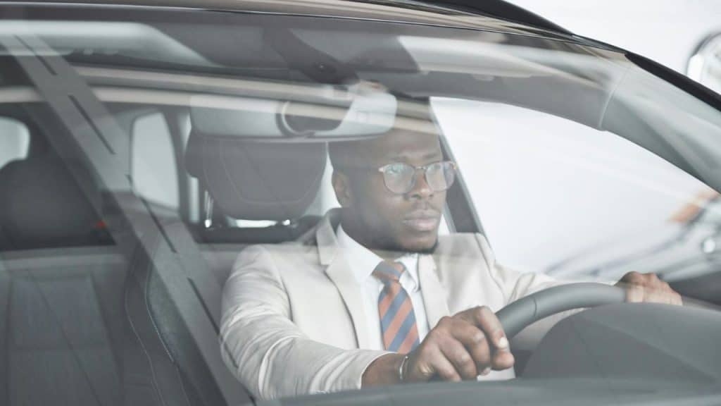 A man in a light suit and glasses sits behind the steering wheel of a car.