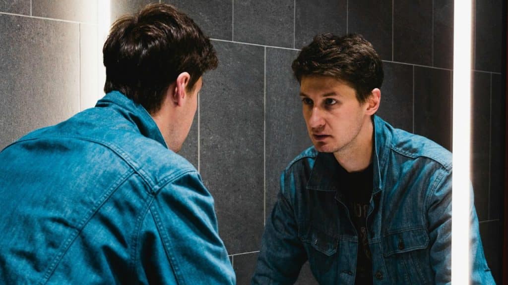 A man in a denim jacket looks intently at his reflection in a bathroom mirror.