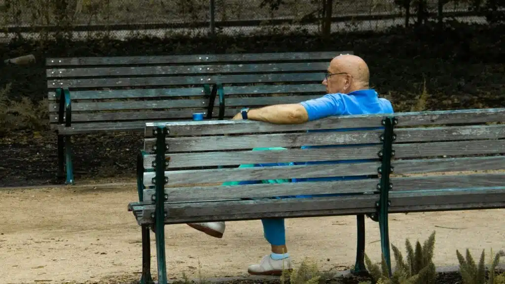 An older, bald man in a blue shirt sits on a wooden park bench outdoors.