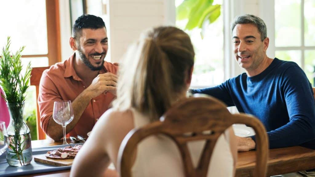 Two smiling men talk to a woman at a dining table in a bright room.