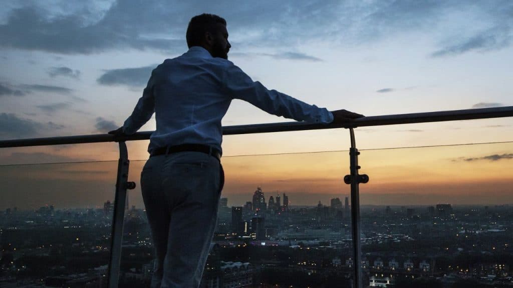 A silhouetted man in a dress shirt stands on a rooftop balcony looking at a city skyline at dusk.