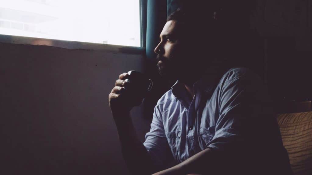 A man sits in a dark room holding a mug, looking out a bright window.