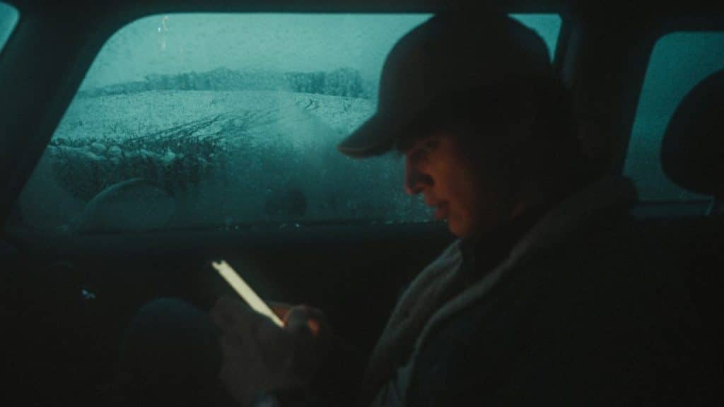 A person in a dark car wears a cap and looks at a glowing phone screen.