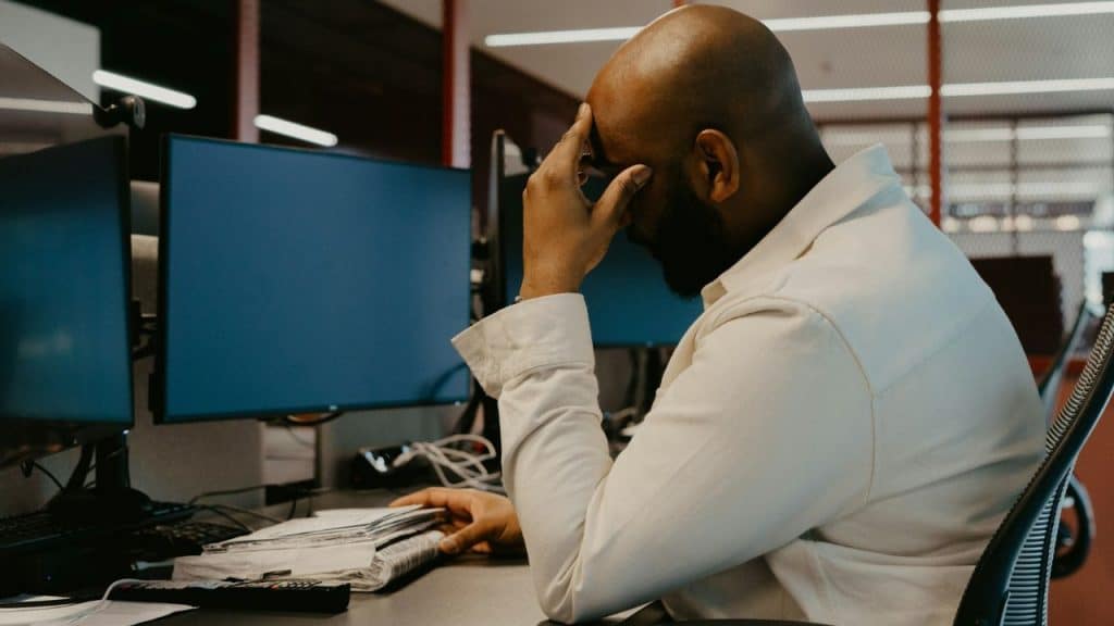 A stressed man sits at a desk with monitors, his head resting in his hand.