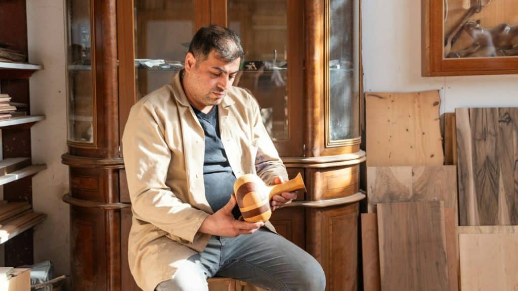 A craftsman in a tan shop coat holds a two-toned wooden mallet in his workshop.