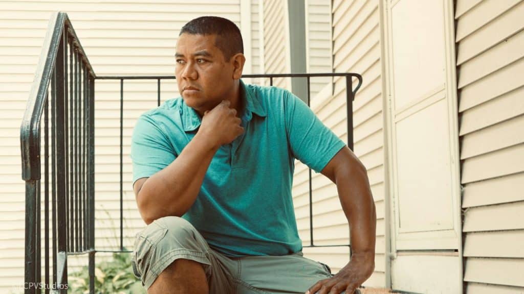 A man in a teal polo shirt sits on a porch step with his hand on his chin.