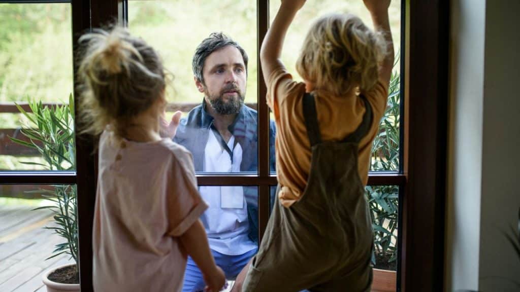 A man looks in through a window at two young children with their backs turned.