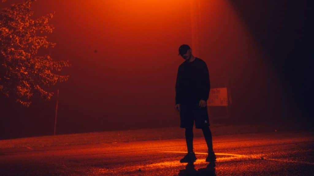 A solitary figure stands on a dark road under bright orange lighting and heavy fog.