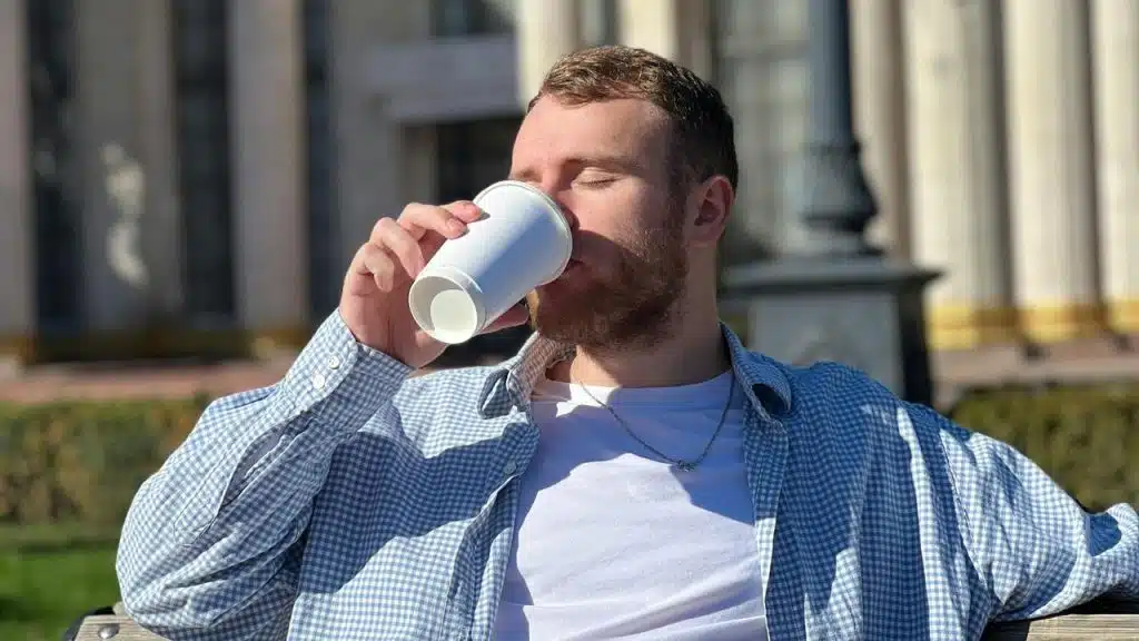 A man sitting outdoors drinking from a paper cup with his eyes closed.