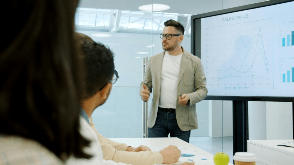 A man in a sport coat gives a presentation with graphs on a large screen to seated colleagues.